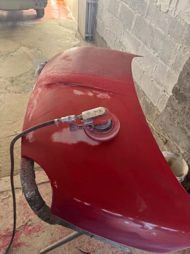 Red car hood being polished with a sanding tool on a workshop table.