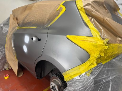 Silver car rear quarter panel partially masked with yellow tape for painting.